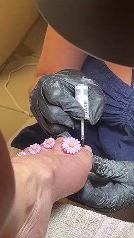 A close-up shot shows a person's foot with small, pink daisy decorations applied to the toenails. Another person, wearing black gloves, is carefully applying a clear substance from a small bottle labeled "BASE" to one of the toenails with a thin applicator. The background is blurry but appears to be an indoor setting, possibly a nail salon, with a lamp positioned overhead. The overall scene depicts a nail beautification process.