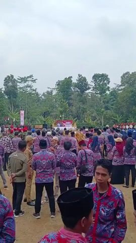 The video shows a crowd of people gathered outdoors, likely for an event. Many individuals are wearing matching shirts with a distinctive red and blue patterned design. Some are wearing tan or khaki-colored shirts. In the background, there appears to be a stage or platform with banners and a gathering of people. Trees and foliage form the backdrop, suggesting a rural or natural setting. The sky is overcast. The focus is on the people in the foreground, some of whom are facing away from the camera, while others are turned towards the camera. The overall impression is of a formal gathering or ceremony taking place in an open area.