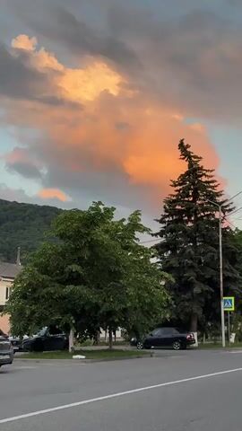 This is a vertical, eye-level video of a street scene at dusk, featuring a dramatic sky with a large, bright orange cloud illuminated by the setting sun. In the foreground, a paved road with white lane markings is visible, with a gray curb running along the side. To the left of the road, there is a sidewalk and a grassy area. A dark green deciduous tree stands prominently on the left, with its branches reaching towards the sky. Behind it, a tall, dark green evergreen tree dominates the center of the frame, its silhouette sharply defined against the colorful sky. Several power lines are strung across the upper portion of the scene. To the right of the evergreen tree, a lamppost stands tall, and a blue and yellow pedestrian crossing sign is mounted on it. A dark-colored sedan is parked on the side of the road, partially obscured by the deciduous tree. Another vehicle, possibly a car, is also parked further down the road to the left. In the background, a tree-covered hill or mountain rises gently under the fading light. The overall impression is one of a tranquil evening scene, with the vibrant sky creating a striking contrast with the darker elements of the landscape.