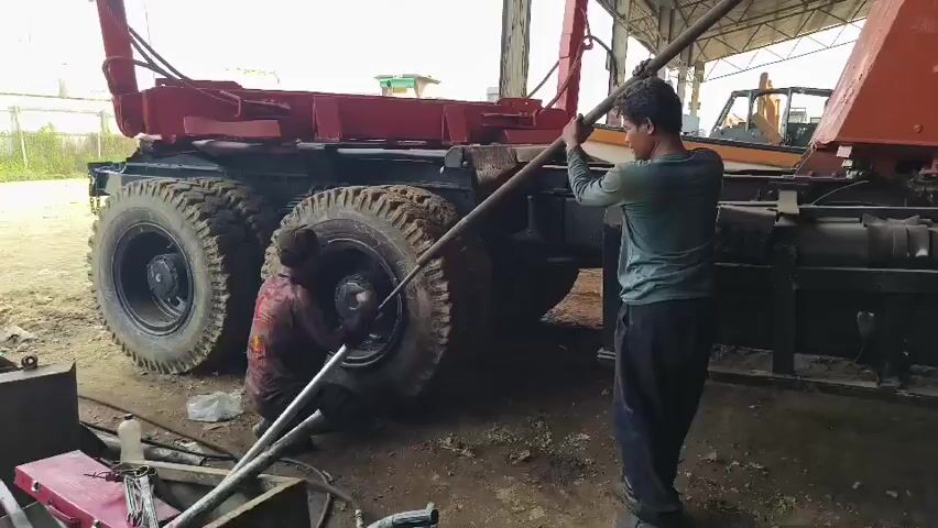 TWO MEN WORK ON THE REAR WHEELS OF A LARGE TRUCK. THE MAN ON THE LEFT IS CROUCHED DOWN AND APPEARS TO BE USING A TOOL ON THE WHEEL HUB. THE MAN ON THE RIGHT IS STANDING AND HOLDING A LONG METAL ROD, WHICH HE IS USING TO LEVER SOMETHING NEAR THE WHEEL. THE TRUCK IS RED AND BLACK AND HAS SEVERAL LARGE TIRES. THE SCENE TAKES PLACE OUTDOORS UNDER A ROOFED STRUCTURE, WITH SOME MACHINERY VISIBLE IN THE BACKGROUND. THE GROUND IS DIRT AND THERE ARE VARIOUS TOOLS AND OBJECTS SCATTERED AROUND. THE LIGHTING IS NATURAL AND APPEARS TO BE DAYTIME.