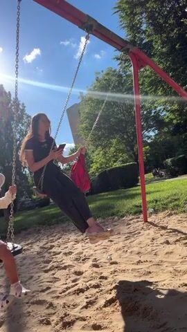 Two females, one adult and one child, are swinging on swings in a sandy playground on a sunny day. The adult female is wearing a black shirt and pants, pink shoes, and is looking at her phone. The child is wearing a white long-sleeved shirt with polka dots, pink shorts, and a white hat with a floral pattern, and is looking around while swinging. A male is briefly visible on the left side of the frame as the camera moves. In the background, there are trees, grass, and a tall building. The sun is shining brightly, creating lens flares in the video.