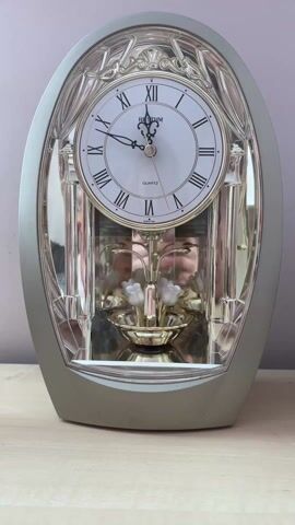 A static shot shows a decorative Rhythm quartz clock, silver and clear plastic, with a white face and Roman numerals. Underneath the clock face is a golden decorative piece with white flowers. The clock rests on a light-colored wooden surface against a pale purple or gray wall.  The reflection of the person operating the camera can be seen on the shiny surfaces of the clock.