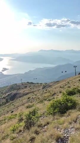 The video opens with a sweeping vista of a sunlit bay or large body of water, surrounded by mountains. The sun is high in the sky, casting a bright glare on the water. In the foreground, a grassy, hilly landscape slopes downwards. As the video progresses, the camera pans to reveal a ski lift or cable car system with several cabins ascending the mountainside. The viewpoint then shifts to a thrilling ride down a mountainside on a roller coaster or toboggan track. A person is seated in the front cart, seemingly controlling the speed. The track winds down the steep, grassy slope, with cable cars visible in the background. The ride offers a panoramic view of the mountains and the water below, creating a sense of speed and adventure.