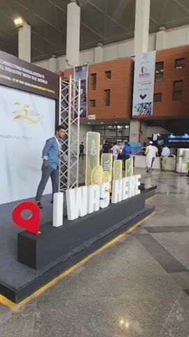 The video shows a man standing on a display stage at what appears to be an exhibition or trade show. He is wearing a light blue button-down shirt and dark pants. To his left is a large sign that reads "25th TEXTILE Series of Exhibitions Bangladesh Edition" and also mentions "25 YEARS OF CONNECTING BANGLADESH'S TEXTILE & APPAREL INDUSTRY WITH THE WORLD." To the right of the man is a large, illuminated sign that says "I WAS HERE" in white block letters. In front of the "I WAS HERE" sign, on a raised black platform, is a red map pin icon. Two black backpacks are placed on the floor to the left of the stage. The background shows a large indoor space with exposed ceiling structures and brick-like wall textures, suggesting a convention center or exhibition hall. Other people can be seen in the background, and there are various displays and signs further into the venue. The man is looking down and appears to be posing for a photo or considering his next move.