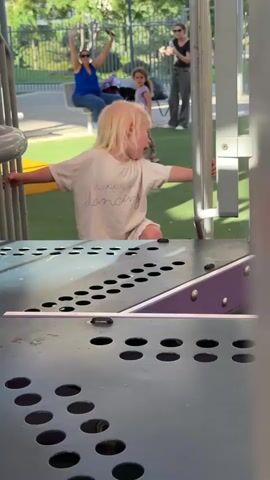 The media file shows a split screen. The top portion features a young child with blonde hair in the foreground, reaching out to a playground structure with their hands. They are wearing a light-colored t-shirt with the text "happy just dancing" embroidered on it, and matching shorts. The child appears focused on their movement.

In the background of the top portion, several other people are visible on what seems to be a playground. A woman in a blue top and another in a dark top are interacting, while a child with dark hair is partially visible. The setting appears to be outdoors, with sunlight suggesting daytime. The presence of playground equipment, green turf, and a fence indicates a recreational area.

The bottom two portions of the split screen show a close-up of a textured, dark gray metal surface with rows of circular holes. These holes are arranged in patterns, possibly part of a musical instrument or a sensory play element on the playground. The lighting on these surfaces suggests the sun is casting shadows.

Overall, the media captures a moment of play and activity on a playground, likely during daylight hours. The focus on the young child and the playground equipment, along with the close-up of the textured surface, creates a sense of engagement and exploration. The location context provided is Petah Tikva, Israel.