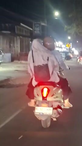 A nighttime shot shows a light-colored scooter with two occupants from behind. The scooter has a license plate that reads "BL 5071 KBK" and the year "12-23". The passenger in the back is wearing a light-colored hijab and a backpack. Both individuals are wearing light-colored jackets. The scooter's red taillight is illuminated, and the scene is lit by streetlights and the lights of other vehicles and buildings in the background. Traffic appears to be moving slowly.