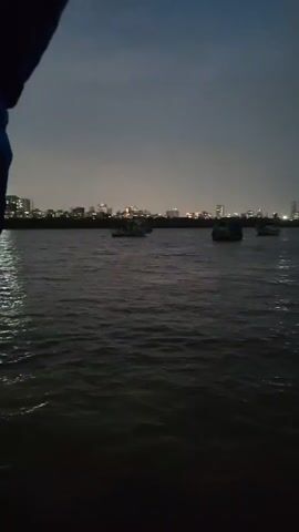 The video shows a body of water with several boats in the distance. The sky is dark and cloudy, suggesting it is either evening or nighttime. In the background, a city skyline with lights is visible. The foreground shows the dark, rippling surface of the water. A portion of a dark-colored garment, possibly a sleeve or shoulder of clothing, is visible in the upper left corner of the frame, indicating a person is recording the scene.