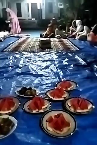 The video shows an outdoor gathering with many people sitting on mats spread over a blue tarp. In the foreground, there are several plates of fruit, including sliced watermelon, bananas, and other round fruits. In the background, there are more mats, people sitting, and a building with lights. One person in a pink hijab is standing and appears to be serving food. Another person in a black outfit is also visible. The lighting suggests it is evening or night.