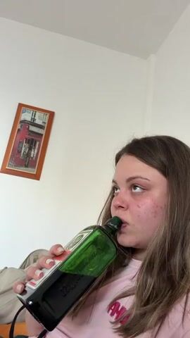 A young woman is lying down, tilting back her head and drinking directly from a green glass bottle. She is wearing a pink t-shirt. The bottle is large and has a black label on the front; it appears to be a bottle of Jägermeister. She has fair skin and is facing the camera. Her long, light brown hair is slightly disheveled. Her face has some acne. The background is a plain white wall with a framed picture hanging on it. The picture seems to be of a building with a red facade. A grey backpack is visible in the lower-left corner of the frame. The lighting is bright. There is ambient background noise with what sounds like a conversation.