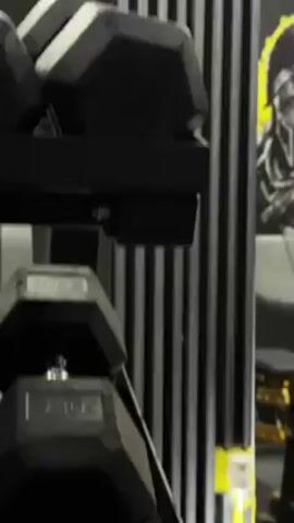 The video shows a black, multi-tiered dumbbell rack with rubber-coated, hexagonal dumbbells on each level. The second tier from the top has a pair of 30-kilogram dumbbells. Behind the rack is a wall with alternating black and gray vertical stripes and a yellow trim near the floor. Part of a poster is visible to the upper right. The camera is stationary.