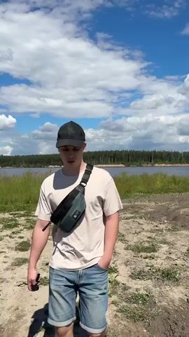 The video opens with a medium shot of a young man walking towards the camera. He is wearing a light-colored t-shirt, blue jeans, a dark baseball cap with a green logo, and a dark green and black fanny pack worn across his chest. His right hand is in his pocket. The background shows a dirt path, green grass, a body of water, and a line of trees under a cloudy blue sky. As the video progresses, the camera zooms in on the man's face. He stops walking and looks directly at the camera with a slight smile, his mouth slightly open as if speaking. The fanny pack and the lower part of his face and shoulders are clearly visible. The background remains the same.