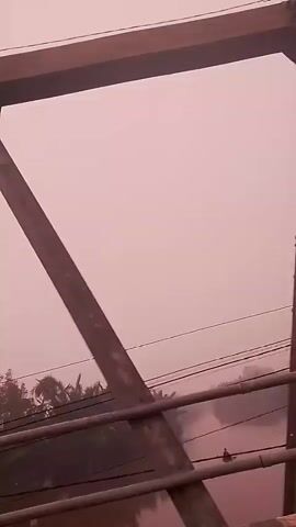 The video shows a view from a bridge, looking downwards. A large metal structure, part of the bridge, dominates the foreground. Below the bridge, there is a body of water, possibly a river, with trees and foliage on the banks. Several electrical wires are also visible, stretched horizontally. The sky appears hazy and pinkish, suggesting a foggy or smoky atmosphere. The overall scene is industrial and somewhat muted in color due to the atmospheric conditions.