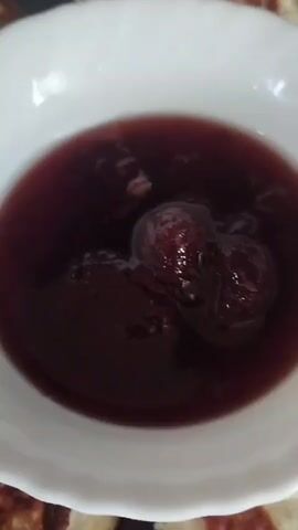 The media file shows a close-up of a white bowl filled with a dark red jam or sauce containing whole berries, possibly cherries or plums. The bowl is placed on a dark surface, possibly a table, and is surrounded by a plate of golden-brown, circular fritters or pancakes. The fritters appear to be lightly browned and have a slightly uneven texture. The lighting suggests it might be indoors, with a soft light illuminating the food. The focus is primarily on the food presentation, emphasizing the rich color of the jam and the texture of the fritters. There is no visible text in the media file. The setting appears to be a domestic environment, likely a kitchen or dining area in Horenka, Ukraine, given the provided location context. The scene evokes a sense of comfort and perhaps a homemade meal, with the jam and fritters suggesting a sweet or savory treat. The overall mood is peaceful and appetizing.