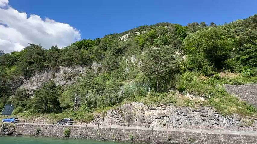 A scenic view of a steep, tree-covered mountainside adjacent to a road and a body of water. The road is bordered by a stone wall, and a small black car is visible driving along it. Further along the road, a stone wall also borders the water, and a pier with steps leading into the water is visible. A large white boat with red trim is docked at the pier, and several people are visible on the pier and on the boat. The sky is clear and blue with a few white clouds. The sun is shining, casting shadows on the mountainside.