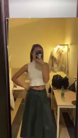 A woman is taking a selfie in a mirror. She is wearing a white, sleeveless, turtleneck top and dark green pants. Her hair is pulled back. She is holding a black and white phone in her right hand. The room has a bed, a desk, and a coat rack with a white fur coat. A black duffel bag is on the desk next to a water bottle. The room is lit by a lamp.