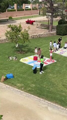The video shows a group of children playing on three large game boards placed on a grassy lawn. The game boards have a pattern of colored squares. There are also a few adults present, seemingly supervising the children. One woman appears to be directing the games and another man stands near one of the game boards with a young girl. A large red dice is on the lawn near one of the game boards. The background features well-maintained lawns, trees, and what appears to be a building in the distance.