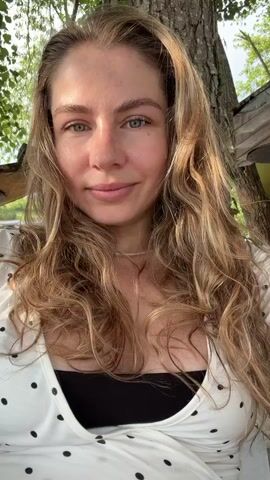 A woman with long, wavy blonde hair and green eyes smiles at the camera. She is wearing a white top with black polka dots and a black undergarment. She appears to be outdoors, with trees and greenery in the background. The sun casts dappled light on her face.