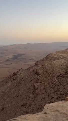 The video captures a wide shot of a vast, rugged desert landscape, likely the Ramon Crater in Mitzpe Ramon, Israel, at either sunrise or sunset. The sky displays a gentle gradient from pale blue at the top to soft orange and pink hues near the horizon, indicating early morning or late evening light.

In the foreground, a steep, rocky mountainside covered in loose scree and larger rock formations slopes downwards. Several individuals, identifiable as hikers, are visible making their way down a winding, narrow path on this slope. Some of these hikers are wearing bright orange or red clothing, making them stand out against the earthy tones of the terrain.

Below the foreground slope, the expansive floor of a large crater or valley stretches out into the distance. A faint, winding line, possibly a road or a dry riverbed, can be seen traversing the valley floor. Beyond the valley, a chain of mountains or hills forms the distant horizon, blending into the soft colors of the sky. The overall scene conveys a sense of grandeur and natural wilderness.