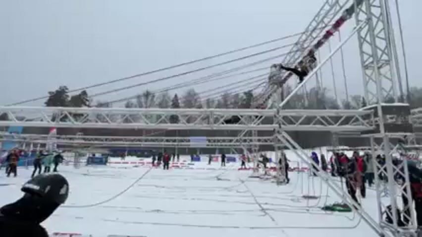 The video shows an outdoor obstacle course challenge set up on a snowy field. There's a complex structure made of interconnected metal beams and ropes stretching across the area. Several individuals are positioned on and around the structure, navigating various obstacles. In the foreground, a person wearing a black helmet and carrying a rope is partially visible. The ground is covered in snow, and there are several people watching the activity in the background. There is also trees in the background. The overall atmosphere appears to be that of a competitive outdoor event.