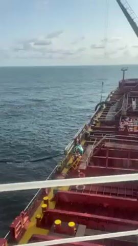 From the deck of a large red ship, the camera captures a view of the open ocean under a partly cloudy sky. The water is dark blue and choppy. The ship's deck is visible, with a complex of red and yellow structures. A crane is situated on the ship. Two people are on the deck, working near some yellow railings and equipment. A dark hose or pipe extends from the ship into the water.