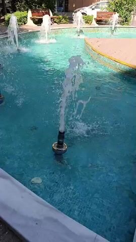 The video shows a close-up of a fountain with multiple jets of water spraying into a bright turquoise pool. The fountain is located in an outdoor urban setting with buildings and trees visible in the background. The water jets are white and foamy, creating a dynamic visual effect. Some of the jets are higher and more powerful than others. The pool has a tiled edge and a wooden decking area at the far end. The overall scene suggests a pleasant and refreshing environment.