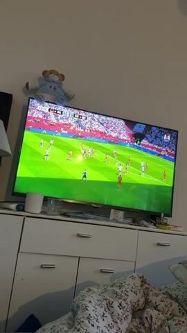 The video shows a television screen displaying a soccer match. The game is being played on a bright green field with players in white and red uniforms. The stands are filled with spectators wearing red. The score on the screen reads 0-0. The television is placed on top of a white dresser with several drawers. On top of the dresser, to the left of the television, is a stuffed teddy bear wearing a blue and white outfit. To the right of the television, a lamp is visible. In the foreground, a person is lying in bed with a blanket patterned with red and white flowers. The scene is indoors, suggesting a living room or bedroom.