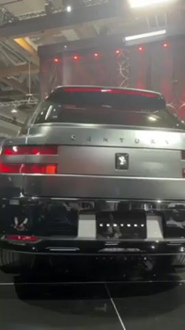 The video shows the rear of a grey SUV with the word "CENTURY" across the back. It is on display, presumably in an indoor setting, as indicated by the lights above and the backdrop of patterned fabric with red lights. The car is in pristine condition, possibly a new model or concept vehicle, based on its appearance and the exhibition setting. There are hints of other vehicles or elements of the display in the background, including a glimpse of a red car interior when the camera shifts slightly. The focus is clearly on the rear design of the "CENTURY" SUV.