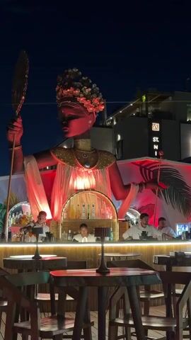A nighttime exterior shot of an outdoor bar area. A large, stylized statue of a woman in elaborate headdress and attire, illuminated in red, dominates the background. In front of the statue is a bar with several bartenders working behind it. Two patrons are visible at the bar, one holding a drink. Tables and chairs are in the foreground. The environment appears festive and possibly tropical, with palm leaf motifs visible.