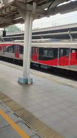A train with red and silver cars is moving from left to right across the frame, passing by a train station platform. The train cars have red doors and windows. The platform is made of grey tiles with a yellow tactile warning strip along the edge. A white pillar supporting the station roof is visible in the foreground. The sky is overcast.