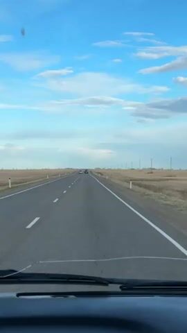 The video depicts a drive along a straight, paved road under a partly cloudy sky. The road has white lane markers and the surrounding landscape is largely flat, dry, and agricultural, with sporadic utility poles dotting the horizon. The view is from inside a vehicle, with the dashboard visible at the bottom of the frame. There are other cars ahead on the road. The video includes music.