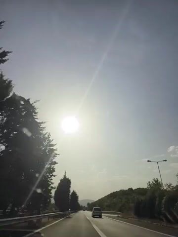 The video shows a view from inside a vehicle traveling on a highway. The sun is bright and appears high in the sky, creating a glare. Trees line the left side of the road, and rolling hills covered in vegetation are visible on the right. Another white vehicle is ahead on the same highway. The sky is mostly clear with a few faint clouds.