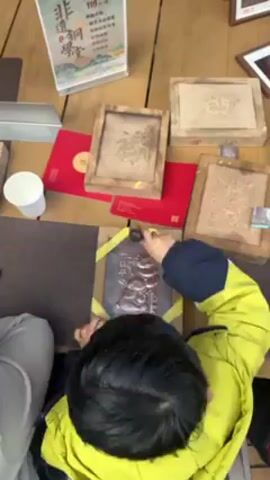 From an overhead angle, a child is seen working on a craft project at a table. The child, wearing a blue jacket with bright yellow accents, is using a small mallet to tap a tool against a dark-colored, framed plaque. The plaque seems to have an image embossed on it. Several other framed plaques are also on the table, along with small red envelopes and a paper cup. A sign with Chinese characters stands upright on the table. The child's activity suggests they are participating in a craft activity, possibly related to Chinese culture. The child's back is to the camera. Their hands are holding the mallet and tool. The workspace is well-lit.