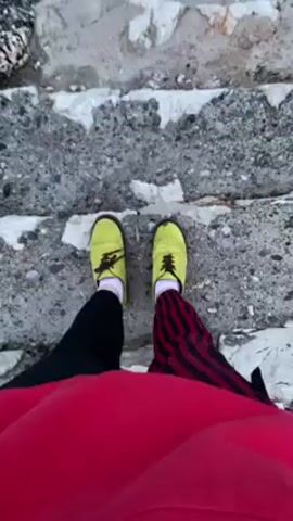 The video shows a person descending a set of weathered, concrete steps leading to a rocky coastline. The person is wearing yellow shoes, black pants on one leg and striped red and black pants on the other, with a red shirt or jacket. As they descend, the camera angle changes to reveal the ocean. The sea is visible, with waves crashing against the dark, rocky shore. The sky is overcast and a muted blue.