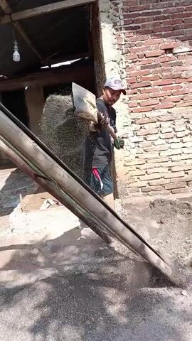 A man wearing a black t-shirt, blue and red shorts, a blue cap, and gloves is shown in an outdoor setting. He is holding a curved blade and appears to be clearing debris from a pile of dirt and gravel. He then picks up a shovel and begins to dig into the pile. The background consists of a brick wall and a partially constructed building with a corrugated metal roof.