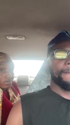 This media file appears to be a short video or a series of still frames captured inside a vehicle, likely a car. The primary subjects are two individuals, a man and a woman, seated next to each other in the front seats.

The man, in the foreground and on the right side of the frame, is wearing a dark sleeveless shirt and a dark blue head covering that resembles a kufi or doek. He also has on rectangular glasses with yellow-tinted lenses and a beard.

The woman, seated to the left of the man, is wearing a maroon top and a patterned headscarf. The headscarf has a repeating pattern that resembles the Gucci logo, featuring interlinked "G"s. Her top also has striped detailing on the sleeves.

The setting is clearly the interior of a car. The dashboard and ceiling of the vehicle are visible, along with the car's interior light. The background shows what appears to be the exterior of the car through the windows, with bright light suggesting it is daytime. There are hints of a grey fabric or object behind the woman.

The activity appears to be a casual moment captured while traveling. The woman is looking towards the camera and seems to be interacting with it, at times smiling. The man is looking forward and slightly to the side. The overall mood seems relaxed and informal.

No explicit text is visible within the frame. The location context provided places the scene in Kwali, Nigeria, suggesting a Nigerian setting for this automotive journey. The bright light implies it's likely daytime, and the weather appears clear.
