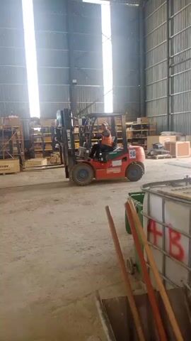 The video shows a forklift driver operating a red and orange forklift inside a large warehouse. The driver is wearing an orange high-visibility vest and a hard hat. The forklift has the number 56 printed on its side. The warehouse appears to be storing various items on shelves. The forklift is moving and its forks are slightly raised. The floor is dirt or concrete. The warehouse has a metal roof and large windows that allow sunlight to enter.