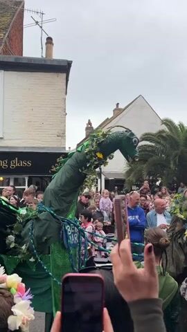 The video captures a street scene during a parade or festival. A large, green dragon puppet is the central focus, surrounded by a crowd of people. Many in the crowd are taking pictures or videos with their phones. The puppet is decorated with greenery and flowers, and seems to be operated by people hidden underneath the fabric.  Other participants in the parade are visible, including one wearing a traditional costume. The background includes buildings with a mix of architectural styles, and a palm tree, suggesting a coastal or temperate climate. The sky is overcast, indicating a cloudy day.  The general atmosphere appears festive and celebratory.