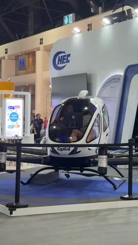The video shows a white eVTOL aircraft on display at what appears to be an exhibition. The aircraft is enclosed in a glass cockpit with two people sitting inside, looking at a laptop. The aircraft is branded with "Capital" on the front and "HEC" on a large blue logo behind it. The background shows exhibition booths with signage, including numbers 31 and 32, and some people walking around. The aircraft is mounted on a stand with black supports and is on a blue carpeted display area.