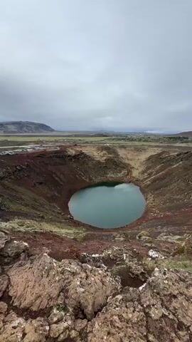 The video shows a crater lake, likely in Iceland, on a cloudy day. The crater walls are reddish-brown and have patches of green vegetation. A path or structure is visible along part of the crater rim. The water in the lake is a muted teal color. The foreground is composed of rocky terrain. In the distance, a flat landscape stretches out under the overcast sky.