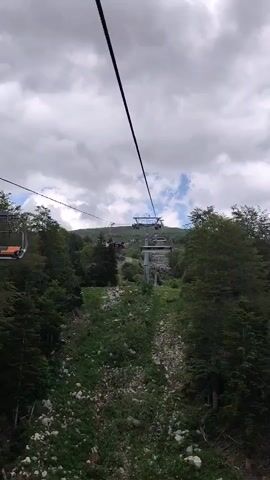 The camera is mounted on a ski lift, providing an upward-looking perspective as it ascends a green mountain slope. Below, a steep, rocky, and heavily vegetated path is visible, flanked on both sides by dense green trees. In the mid-distance and background, several metal ski lift support pylons and multiple individual ski lift chairs are seen continuing up the slope. Some distant chairs appear to be occupied by indistinct figures. The sky overhead is partly cloudy with patches of blue.