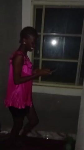 The video shows a dimly lit room. A woman with braided hair is wearing a pink top and black shorts. She is holding a phone and appears to be taking photos or videos. There is a window behind her, and another person is partially visible sitting near a bed or couch. The room looks cluttered. The camera moves around, showing more of the room.