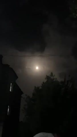 A still, nighttime shot of a full moon partially obscured by clouds. A dark tree line is visible in the lower right corner, and the corner of a dark building is visible in the lower left corner. Power lines are visible stretching across the top of the frame.