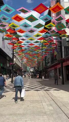 The video displays a vibrant street scene in Andorra la Vella, Andorra, under a clear blue sky with scattered white clouds. The main subject is a pedestrian street lined with multi-story buildings, transformed by an overhead art installation. Hundreds of colorful square fabric decorations, arranged in a grid pattern, stretch across the street above the heads of pedestrians. These squares feature concentric patterns in bright hues including red, orange, yellow, green, blue, and pink, casting interesting shadows on the pavement below.

Numerous people are seen walking casually along the street in both directions, some carrying bags, suggesting a shopping or commercial district. The buildings on either side house various shops and businesses, with display windows and entrances. On the left side, a large banner on a building features an illustration of a woman's face and partially visible text, including "Take care with" and "Andorra." On the right side, a shop sign clearly reads "ROCA FRISA," and another small sign displays "120." The time of day appears to be daytime, with ample sunlight illuminating the scene, contributing to a lively and pleasant atmosphere.
