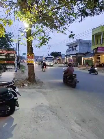 The video shows a street scene at dusk or early dawn. A truck with its headlights on is driving down the road. Several motorcycles are also present, with riders visible on some of them. On the left side of the road, there is a tree with a streetlamp illuminating its leaves. There are also signs and a parked motorcycle visible on the left. Buildings line the right side of the road, some with lights on. The sky is overcast.