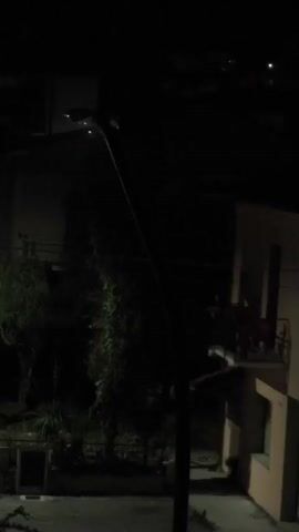 A nighttime exterior shot shows a street lamp and buildings. The street lamp is illuminated and leans slightly to the left. A multi-story building with a balcony and windows is visible on the right. A dark area, possibly containing trees and foliage, is on the left. The scene appears to be quiet and still.