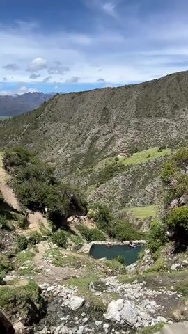 A high-angle, static shot looks down into a narrow, verdant mountain valley. A naturalistic, man-made pool is visible in the center of the frame, filled with turquoise water and occupied by several people swimming and relaxing. The surrounding landscape is characterized by steep, rocky slopes covered in sparse vegetation, with a clear blue sky dotted with white clouds overhead. A winding dirt path is visible on the left side of the valley, leading towards the pool. In the distance, a red-roofed building sits on a patch of green on the hillside. The overall impression is one of a remote, natural, and tranquil setting.