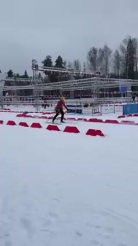 The video shows a biathlon competition on a snowy course marked with red obstacles. Several participants are cross-country skiing through the course. In the background, a large metal structure supports lights and signage, with a building visible beyond. The weather appears overcast.