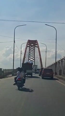 The video shows a view from a vehicle driving on a road, approaching a large, arched bridge. Several vehicles are on the road, including motorcycles, cars, and a truck. The sky is clear with some clouds. The bridge is red and has a distinctive lattice-like structure. There are tall streetlights on either side of the road.