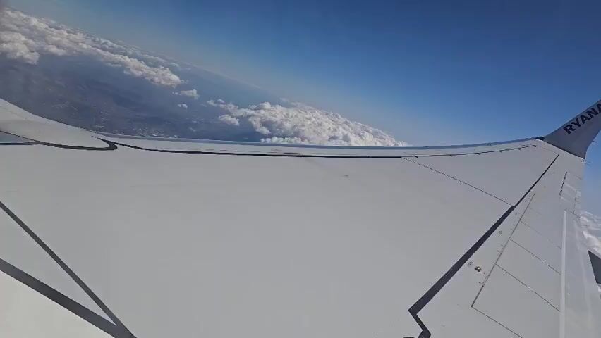 The video shows an aerial view from the wing of an airplane, identified by the "RYANAIR" logo on the wingtip. The plane is flying above a layer of white, fluffy clouds with a clear blue sky above. Below the clouds, a landmass with visible structures suggesting a town or city can be seen in the distance. The angle of the shot is tilted, giving the impression of the plane banking or turning. The lighting suggests it is daytime.