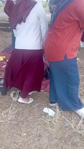 The video shows a group of people, mostly women wearing hijabs, gathered outdoors in what appears to be a park or grassy area. Some are standing, while others are sitting on blankets or the ground. There are trees and foliage in the background, and a white car is parked nearby. One woman is wearing a red top and blue pants, another is in a white sweater and a maroon skirt. A young boy is also visible. There are picnic blankets, bags, and water bottles scattered around. The overall impression is of a casual outdoor gathering.